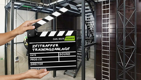 Jetzt Video vom Aufbau der Trainingsanlage ansehen Hände halten Filmklappe, dahinter Metallkonstruktion (Höhensicherungs-Trainingsanlage)