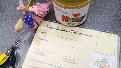 Pädagogik-Projekt mit Bienen-Führerschein Auf einem Tisch liegen Blumen, ein Glas Honig und ein Zettel mit Lanyard