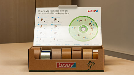 Jetzt umsteigen auf nachhaltiges Verpackungsband. Pappkiste auf einem hellen Tisch, aufgeklappt. In der Kiste sieben Klebebandrollen, Infografik auf der Deckelinnenseite des Kartons.
