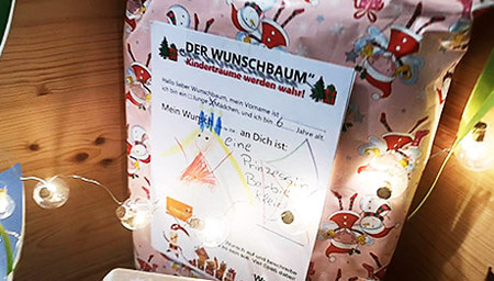Rosa eingepacktes Geschenk mit einem Zettel drauf, der eine Kinderzeichnung einer Prinzessin zeigt und einen Text zum Wunschbaum. Davor eine Lichterkette.