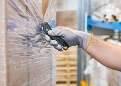 Detail, Verpacktes Gut mit Folie umhüllt, Person, von der Unterarm und Hand in Handschuh zu sehen ist, schneidet mit einem Sicherheitsmesser die Folie ein.