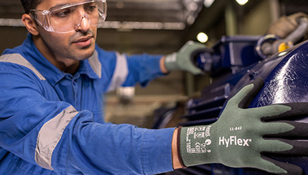 Mann mit blauer Arbeitskleidung, Schutzbrille und grünen Handschuhen arbeitet an einer Anlage