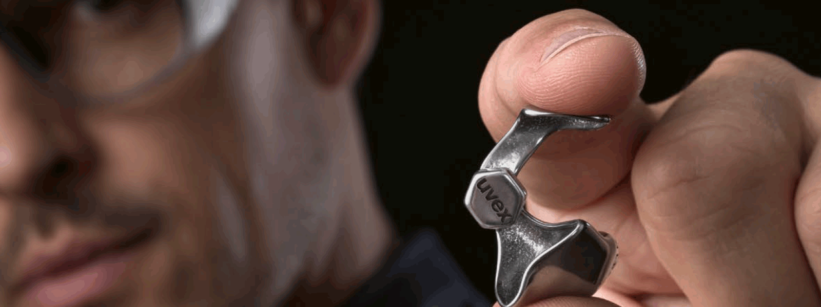 Mann hält eine metallene Otoplastik mit der Aufschrift uvex in die Kamera. Er selbst verschwindet unscharf ins Dunkle