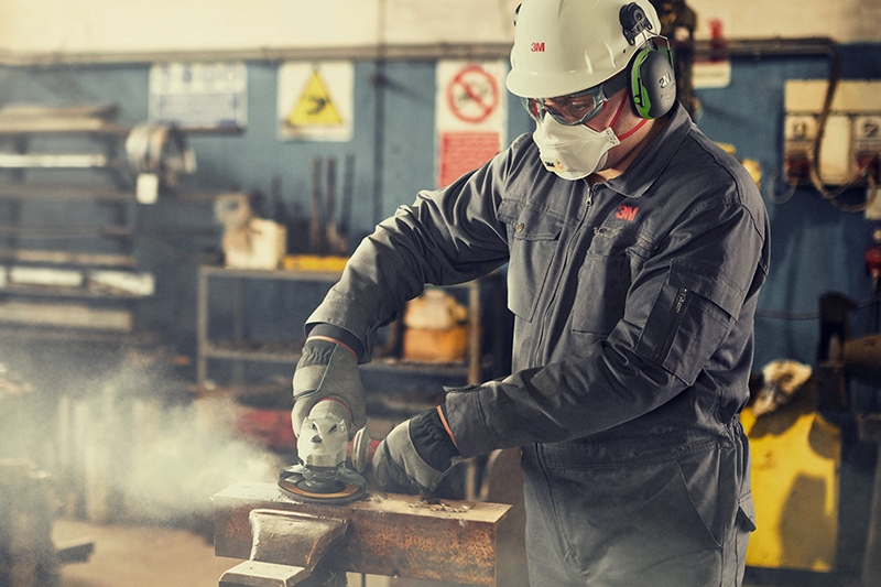 Person mit Mundschutz, Brille, Handschuhen, Arbeitskleidung und Helm in einer Metallwerkstatt beim Flexen