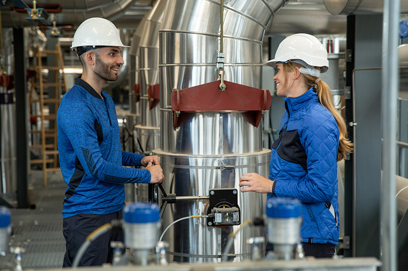 ein Mann und eine Frau in blauer Arbeitskleidung und weißen Helmen in einer Fabrik mit großen Edelstahlrohren